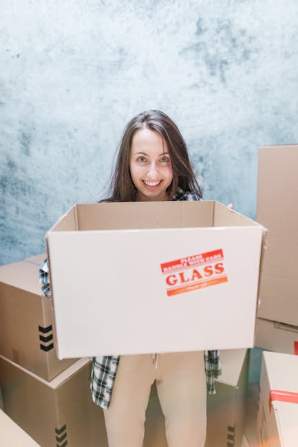 A young woman sitting on a wooden floor inside a residential space, surrounded by numerous cardboard boxes in various sizes, some sealed with red tape and others open. She is wearing casual clothes, i