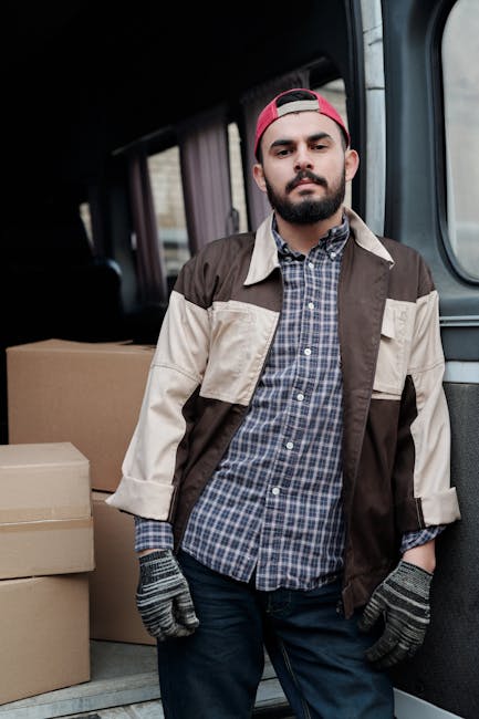 A smiling young man with shoulder-length hair, wearing a bandana and a dark long-sleeve shirt, is sitting in the driver's seat of a white moving van with the words 'MOVING COMPANY LOCAL & LONG DISTANC
