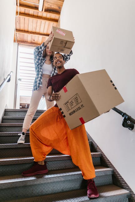 Two movers from Man with Van Finchley are carrying cardboard boxes during a home relocation inside a building with a staircase. The man in the foreground is wearing bright orange trousers, a maroon sh